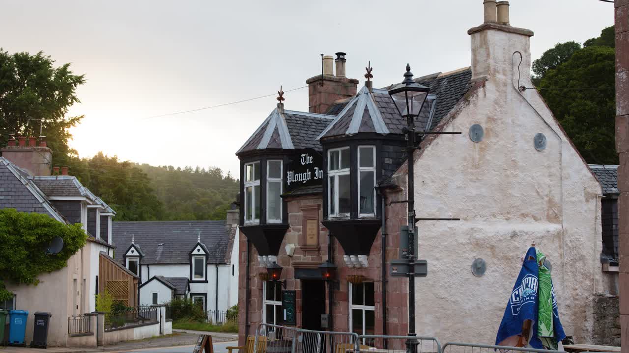 Static wide shot of old stone pub, soft evening light, tranquil village street, minimal movement