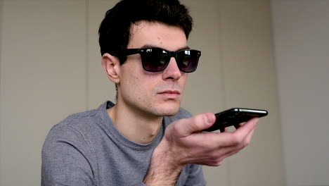 vista de cerca de un hombre ciego con gafas de sol sentado en el sofá de casa mientras sostiene un teléfono inteligente y tiene una llamada de manos libres