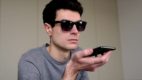 close up view of a blind man in sunglasses sitting on the sofa at home while holding a smartphone and having a hands free call