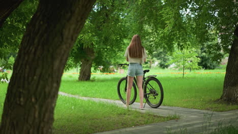 young woman in pink sneakers and jean shorts approaches her bicycle on a tree-lined path, placing an item on the bike seat, background includes lush greenery, a building, and two distant people