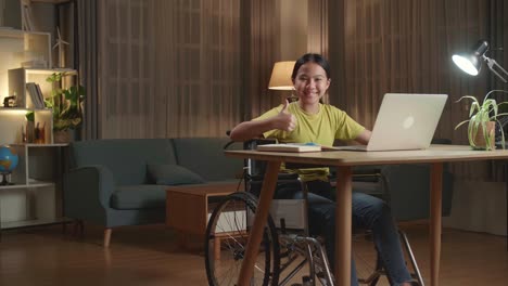 asian kid girl sitting in a wheelchair using laptop computer while smiling and thumb up to camera at home