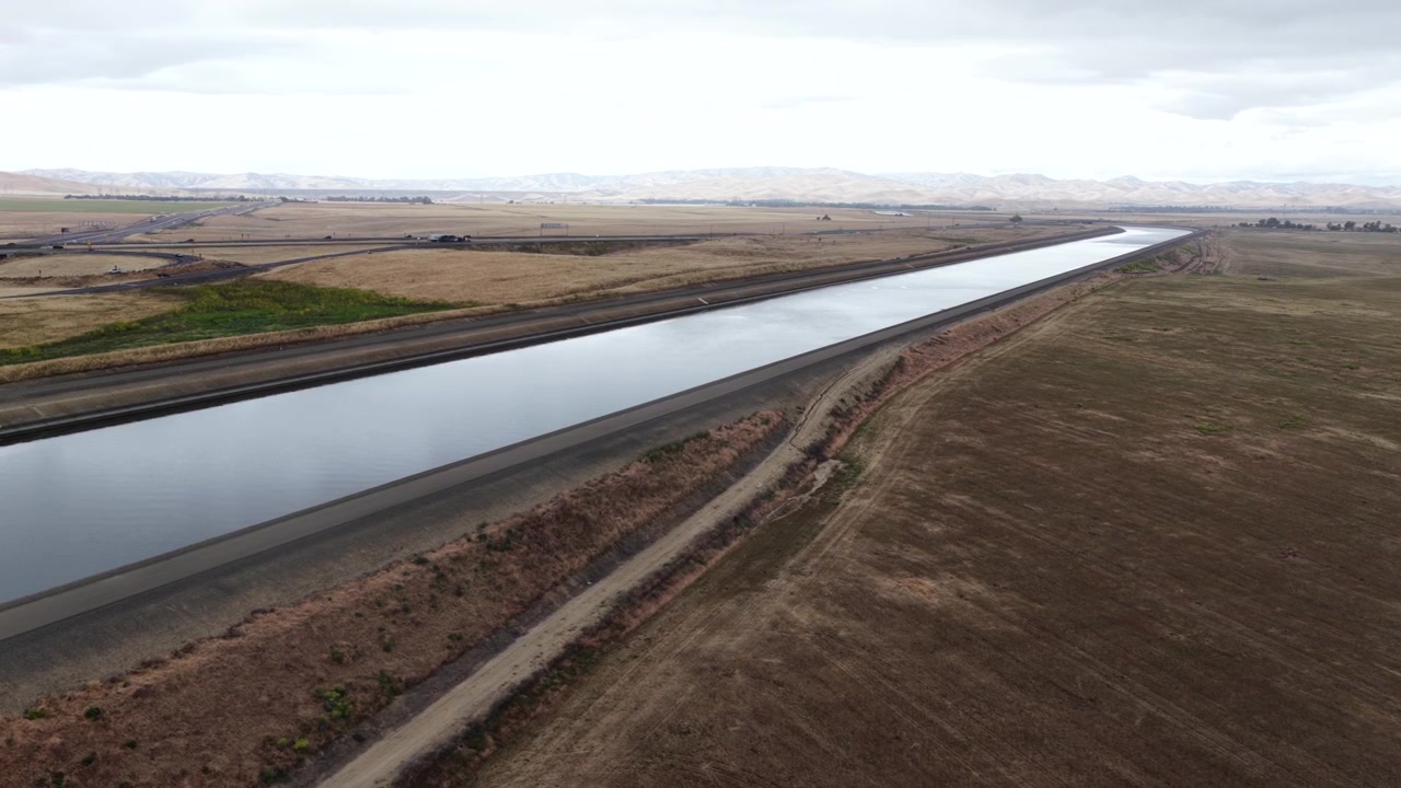 Het Californische Aquaduct loopt recht door droog, vlak landbouwgebied, met een toegangsweg ernaast, voertuigen in de verte en kale bergen die vervagen in een bleke, bewolkte lucht. Californië, VS. Brede luchtopname.