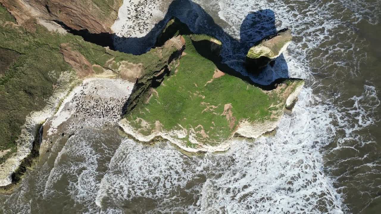 Overhead drone shot captures massive cliff towering over expansive seascape in England, UK. The rugged rock formation, contrasting beautifully with waves crashing it during daytime