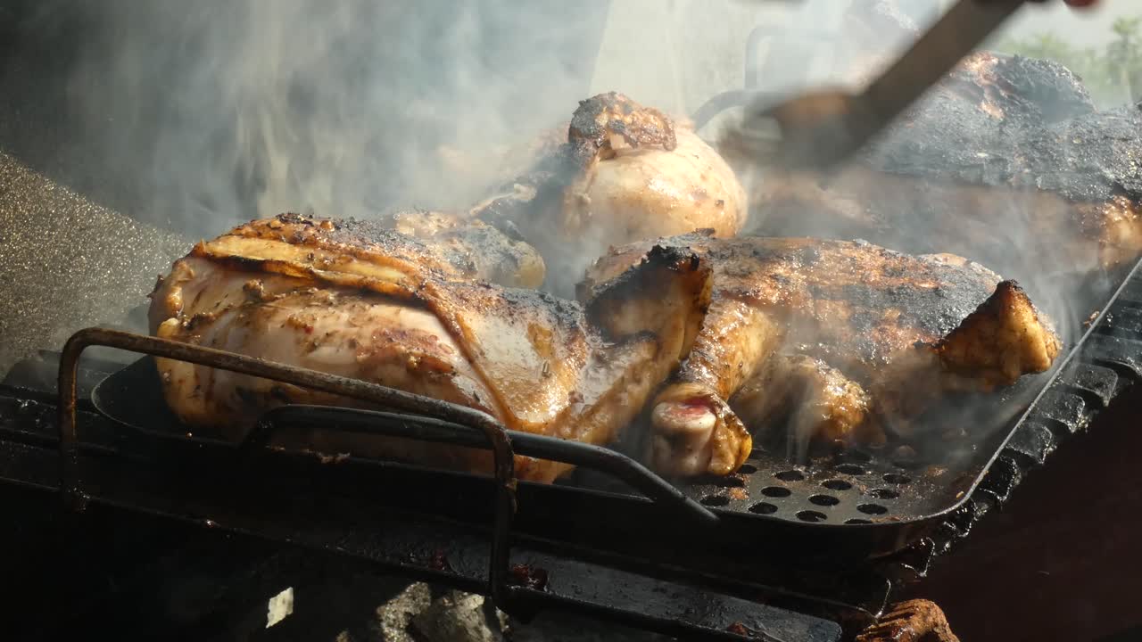 Chicken cooking in a barbecue. A person holding tongs moves the pieces of meat around. Smoke rises from the flames below the bbq grill. Some pieces of chicken appear to be burnt.
