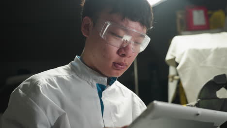 joven estudiante chino en bata blanca de laboratorio se centra en su tableta mientras usa gafas de seguridad transparentes en un taller de automóviles iluminado