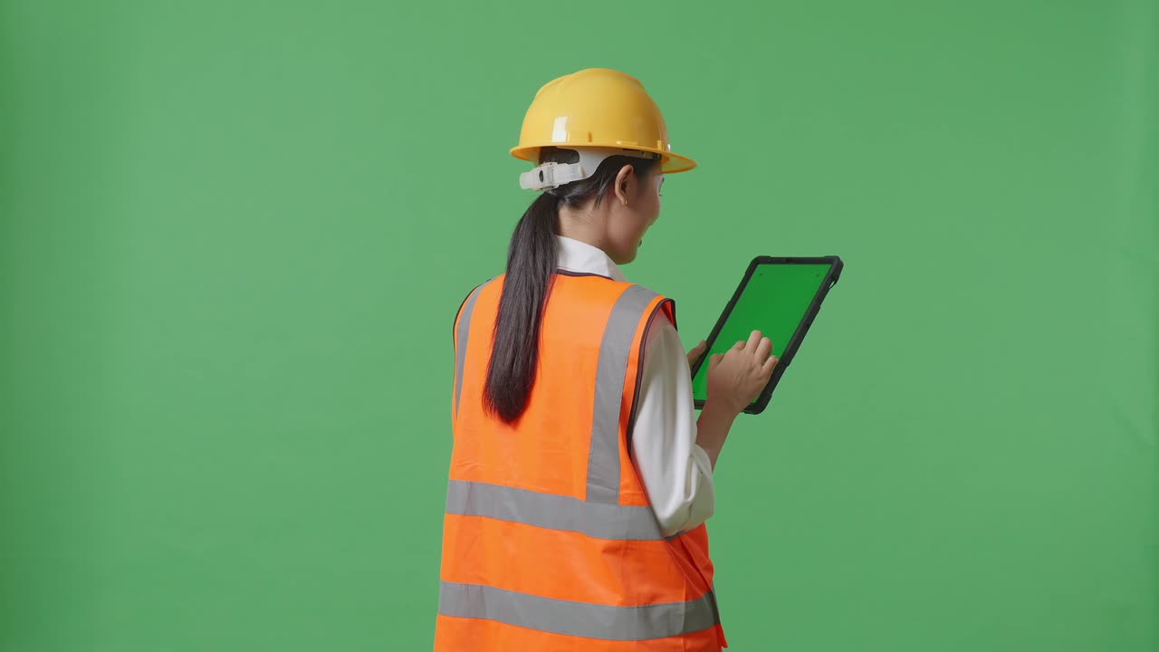Back View Of Asian Female Engineer With Safety Helmet Working On A Green Screen Tablet And Looking Around While Standing In The Green Screen Background Studio