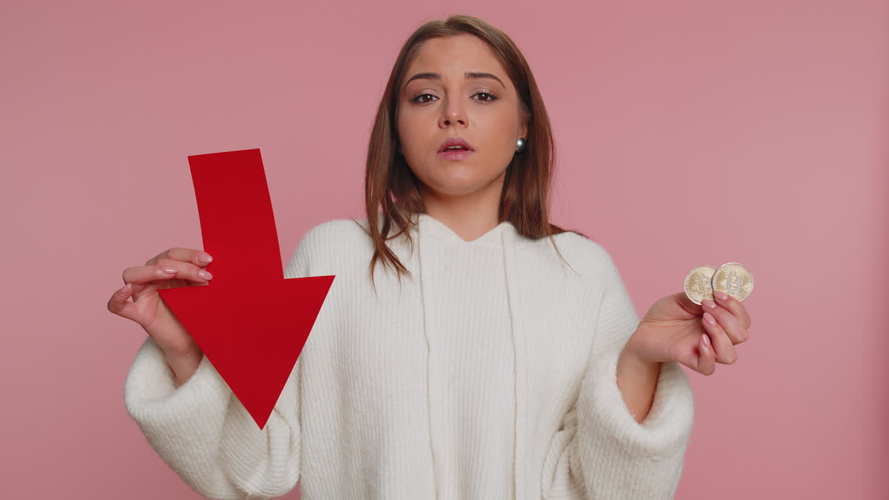 mujer joven mostrando bitcoins dorados y flecha apuntando hacia abajo, minería btc, inversión en criptomonedas