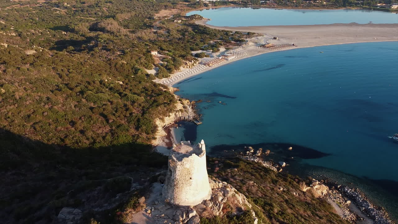 이탈리아 사르데냐의 바위와 관목으로 둘러싸인 토레 디 포르토 지운코 타워의 공중 원형 전망과 관광객들이 랜드마크를 탐험하고 있습니다.