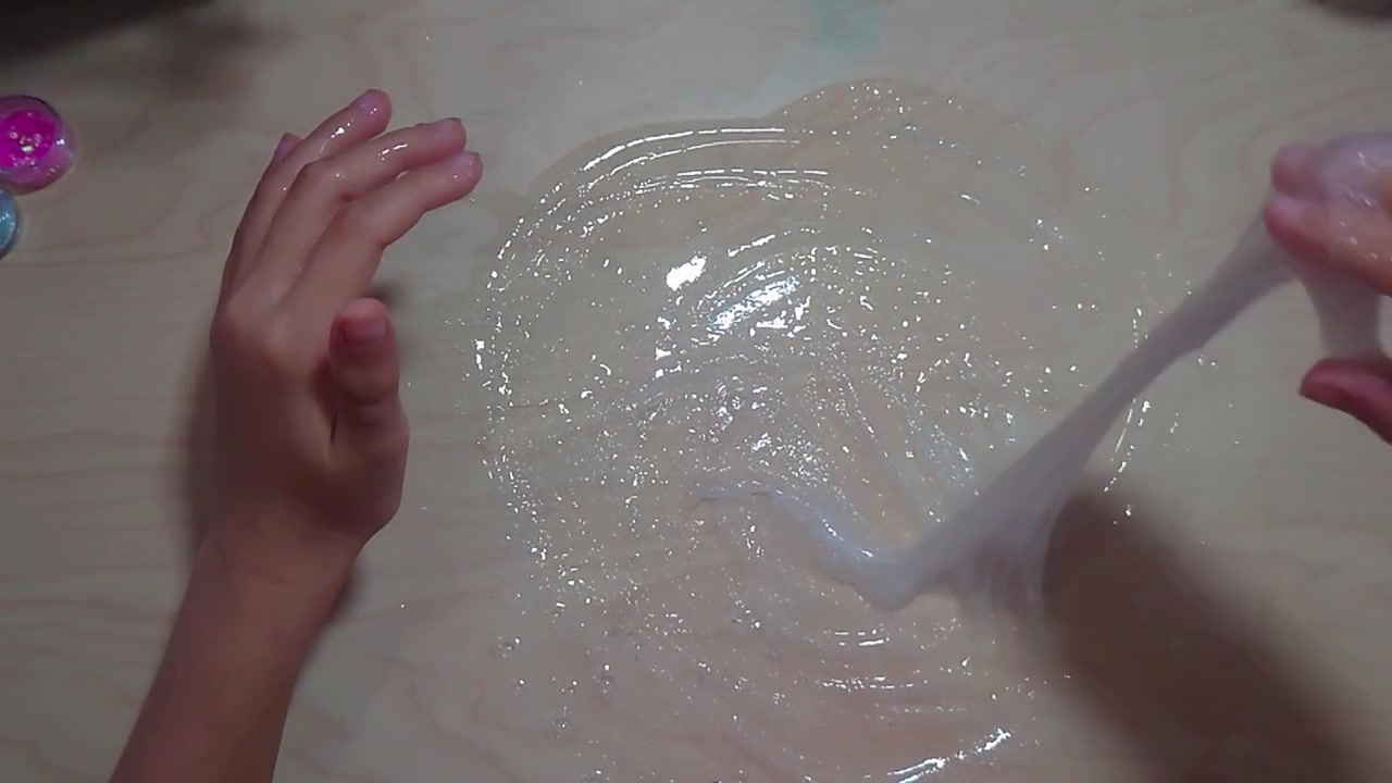 Una ripresa dall'alto mostra un bambino preadolescente che usa entrambe le mani per mescolare una soluzione attivatore liquida in una pozza di colla PVA trasparente su una scrivania di legno liscia durante un progetto artigianale interattivo di scienza domestica.