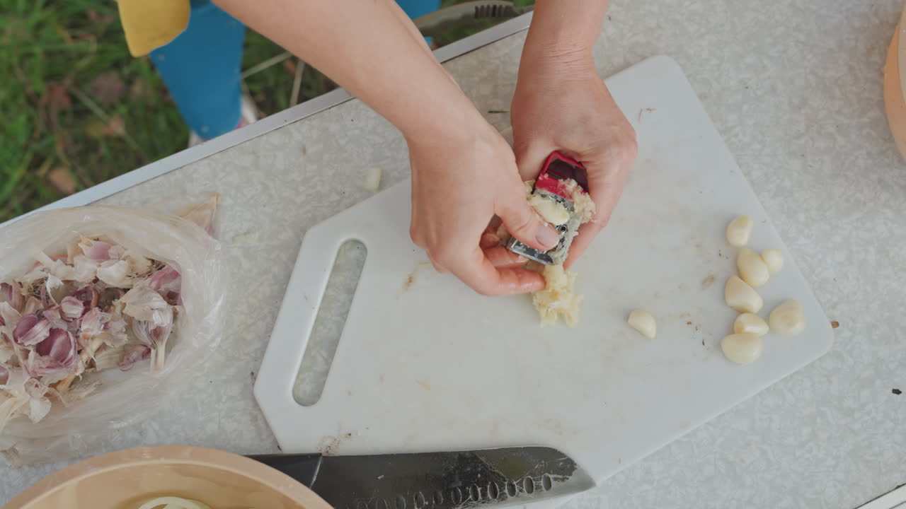 Overhead garlic grating and chopping on board, caucasian camp cook slicing onion and grating cloves at campsite, knife and bowl visible, sunny outdoor table scene for rustic meal preparation