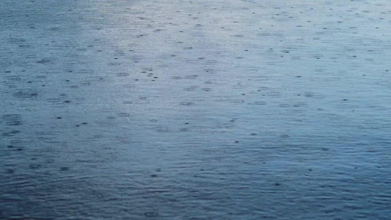 Raindrops fall onto calm blue water surface. Gentle ripples spread outward from each impact. Mood is peaceful, tranquil, and reflective. Perfect for scenes needing calm, natural atmosphere