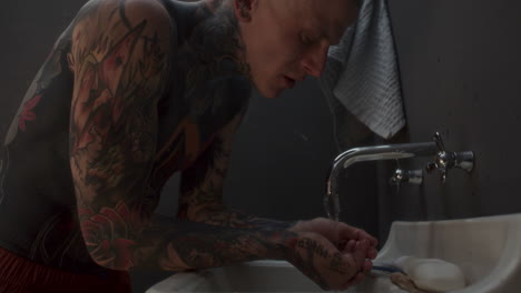 shirtless tattooed man rinsing his face with water above sink in prison cell