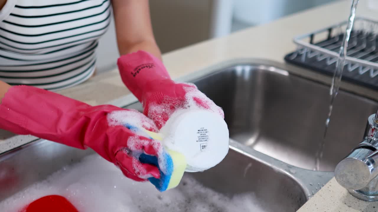 Person wearing gloves washes dishes in a bright kitchen using a sponge and sink