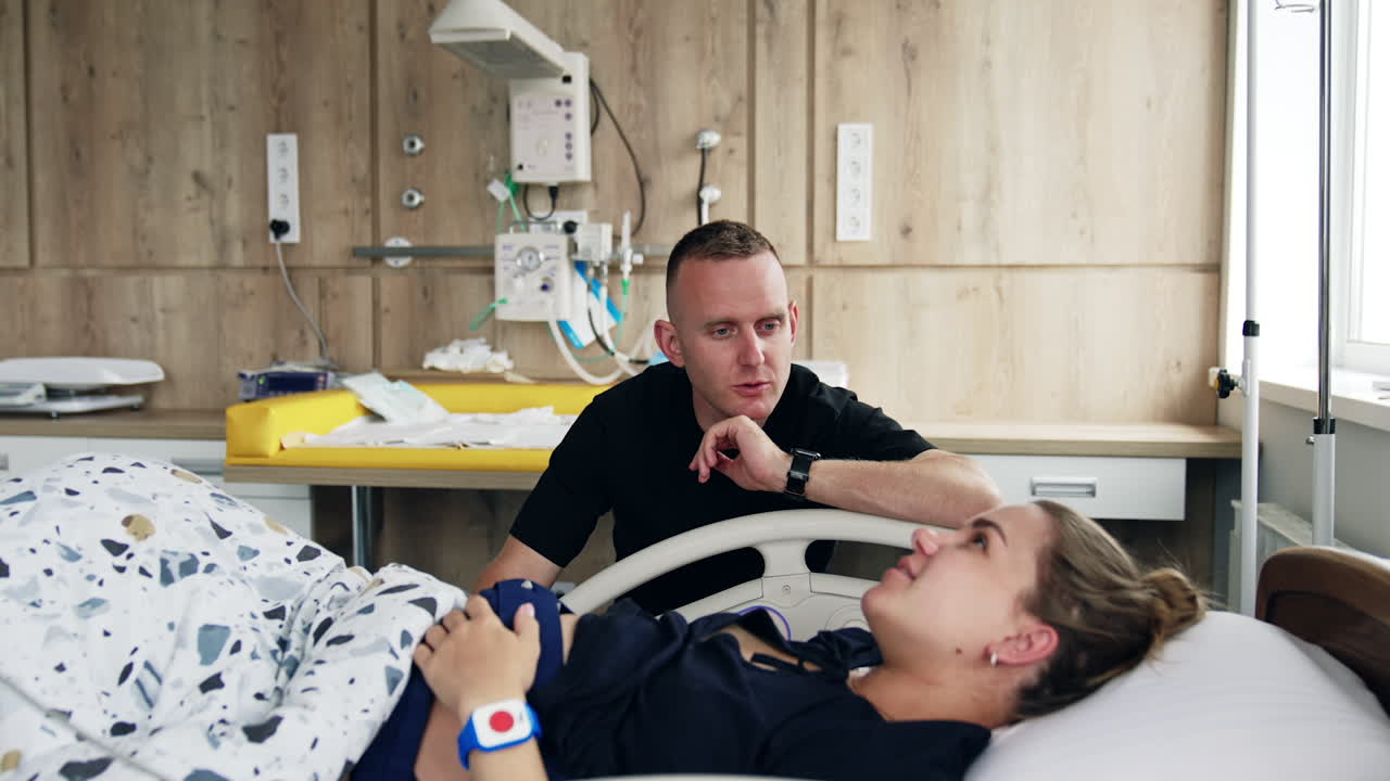 Man wearing black t-shirt talking to his wife lying in hospital bed. Woman is checked before baby delivery.