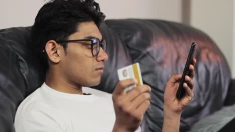 Young-man-customer-holding-credit-card-and-smartphone-sitting-on-couch-at-home