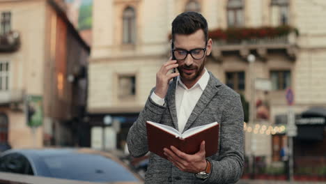 Stylish Businessman In Glasses Standing In The Street, Talking On The Phone And Looking At His Open Planner, Then Going Away