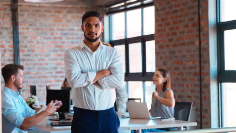 portrait of serious businessman standing in busy office with colleagues working in background