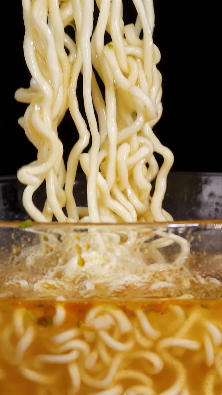 Una mano usa le bacchette per sollevare noodles istantanei cotti da una ciotola di vetro fumante di brodo, ripreso in primo piano con illuminazione drammatica e sfondo minimale.