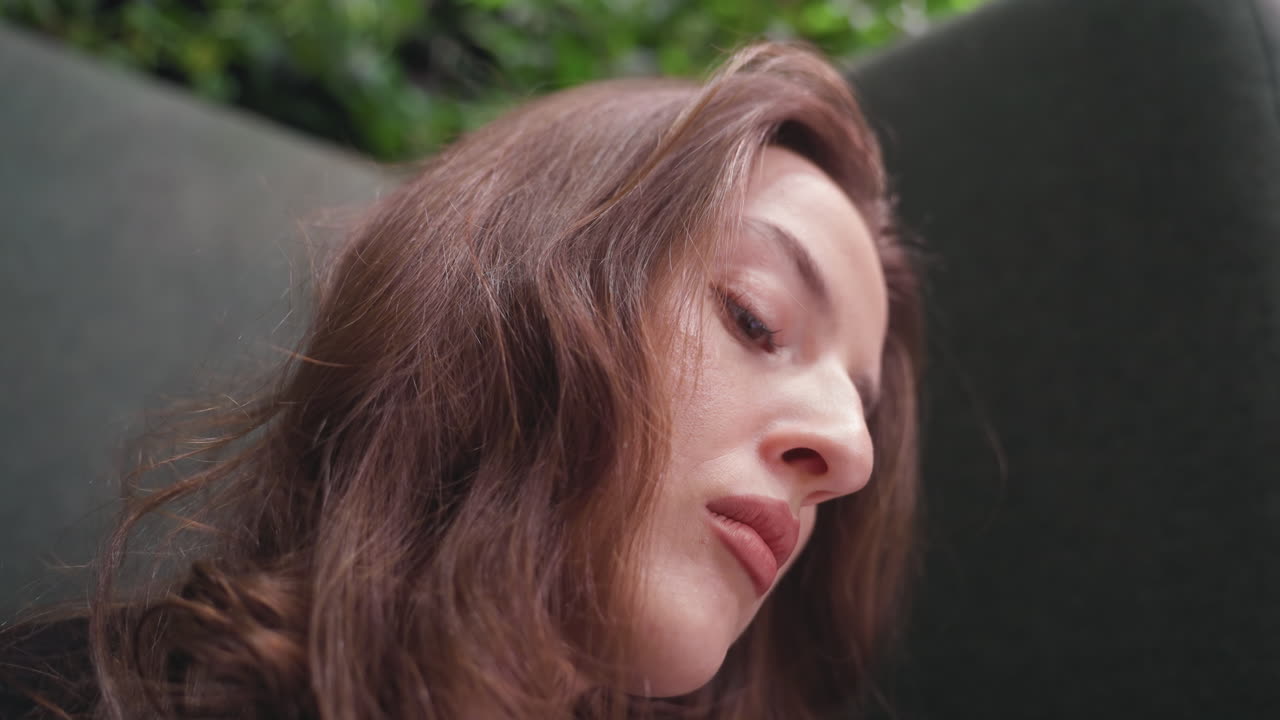 Close up of light skin lady with blond hair looking downward in focused concentration, soft natural light highlighting her facial features, indoor setting with blurred greenery in background