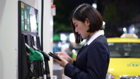 a businesswoman engaged with her mobile device at a fuel station while refueling her vehicle in an evening urban setting