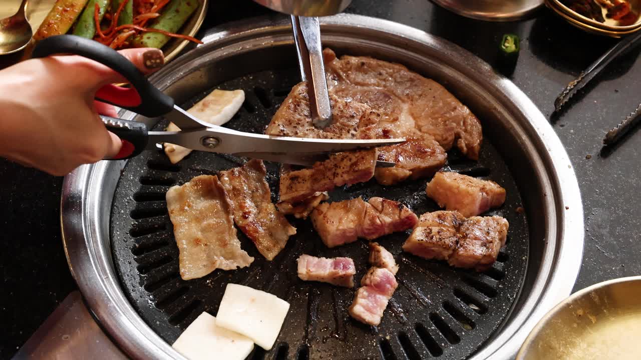 Hand uses scissors to cut pork belly on tabletop grill, bright lighting, overhead close-up view