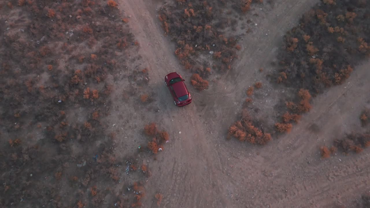 Overhead drone of a red car driving along a dusty desert track at twilight amid dry scrub and forked trails—cinematic travel and off-road establishing shot; empty landscape