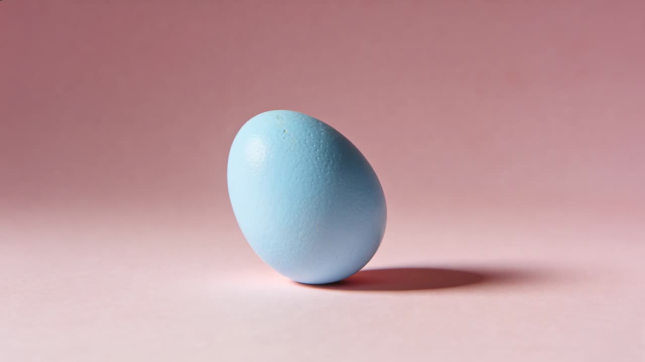Initial tilting matte light-blue egg rolling across pink tabletop settling on side with soft shadow