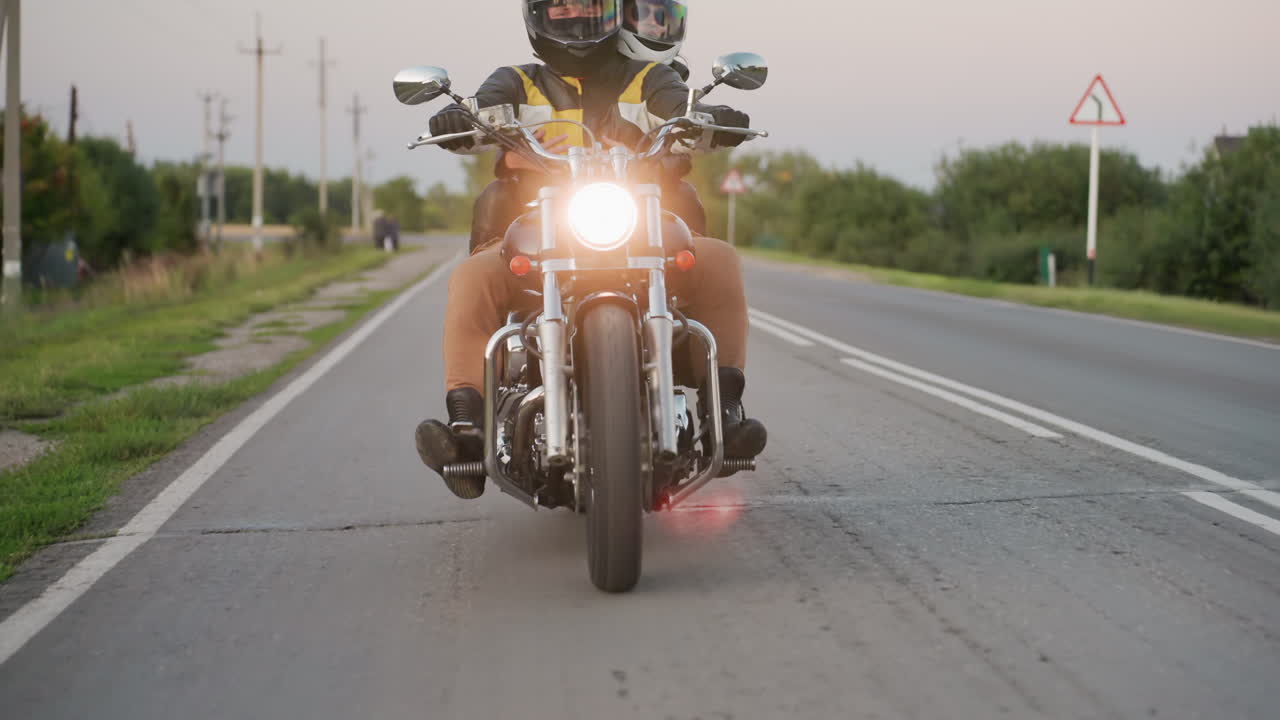 Motorcycle rider with passenger wearing helmets travels down countryside road at dusk, glowing headlight shining on asphalt as trees and roadside vegetation line horizon and sharp bend signpost