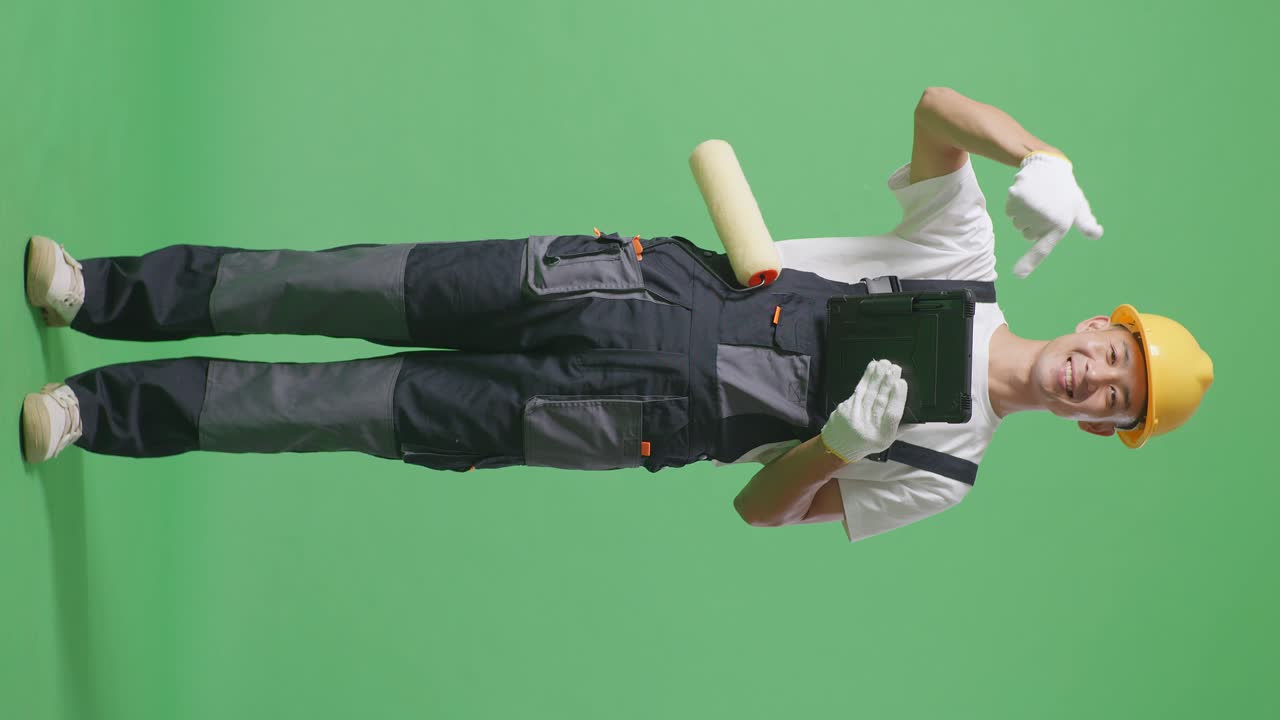 Full Body Of Asian Man Painter Wearing Safety Helmet Smiling And Pointing To A Tablet In His Hand While Standing In The Green Screen Background Studio