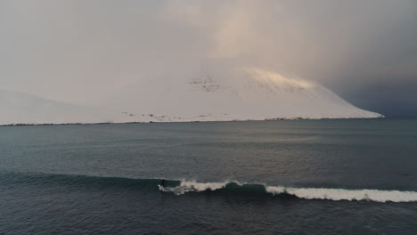 aerial descend to icelandic coastline with crashing waves as surfer takes off pumping and carving along face, water meeting snow covered arctic shores