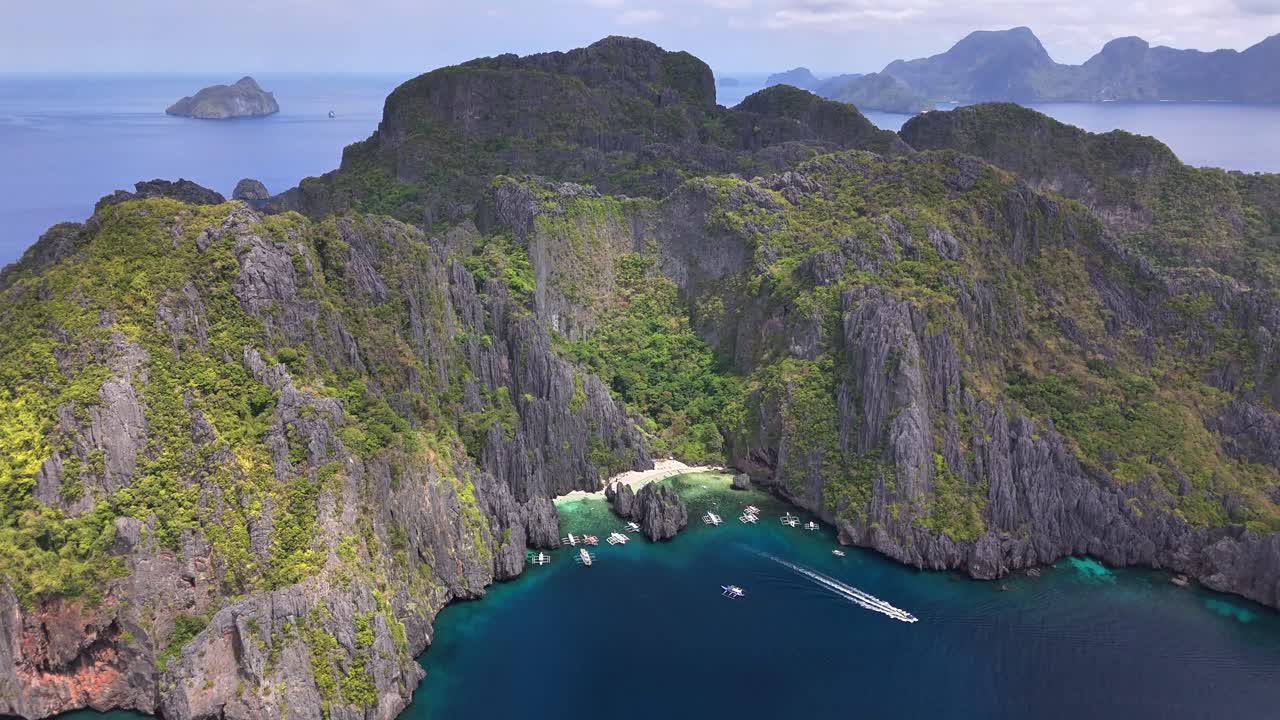 Aerial view of Secret Lagoon Jiji's Beach on Miniloc Island, Philippines, with towering limestone cliffs, vibrant green foliage, turquoise waters, and several boats anchored near the white sandy shore