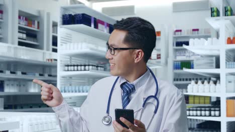 primer plano de un médico asiático con un estetoscopio usando un teléfono inteligente y apuntando hacia un lado mientras está de pie en la farmacia
