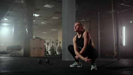 a young slender woman prepares and warms up before training. hitching and stretching muscles after a tedious hard workout in the dark interior of the fitness room