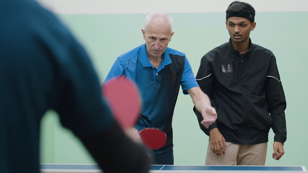 Coach demonstrates table tennis stroke during training session with young athlete while another player watches closely, highlighting mentorship, focus, learning, guidance