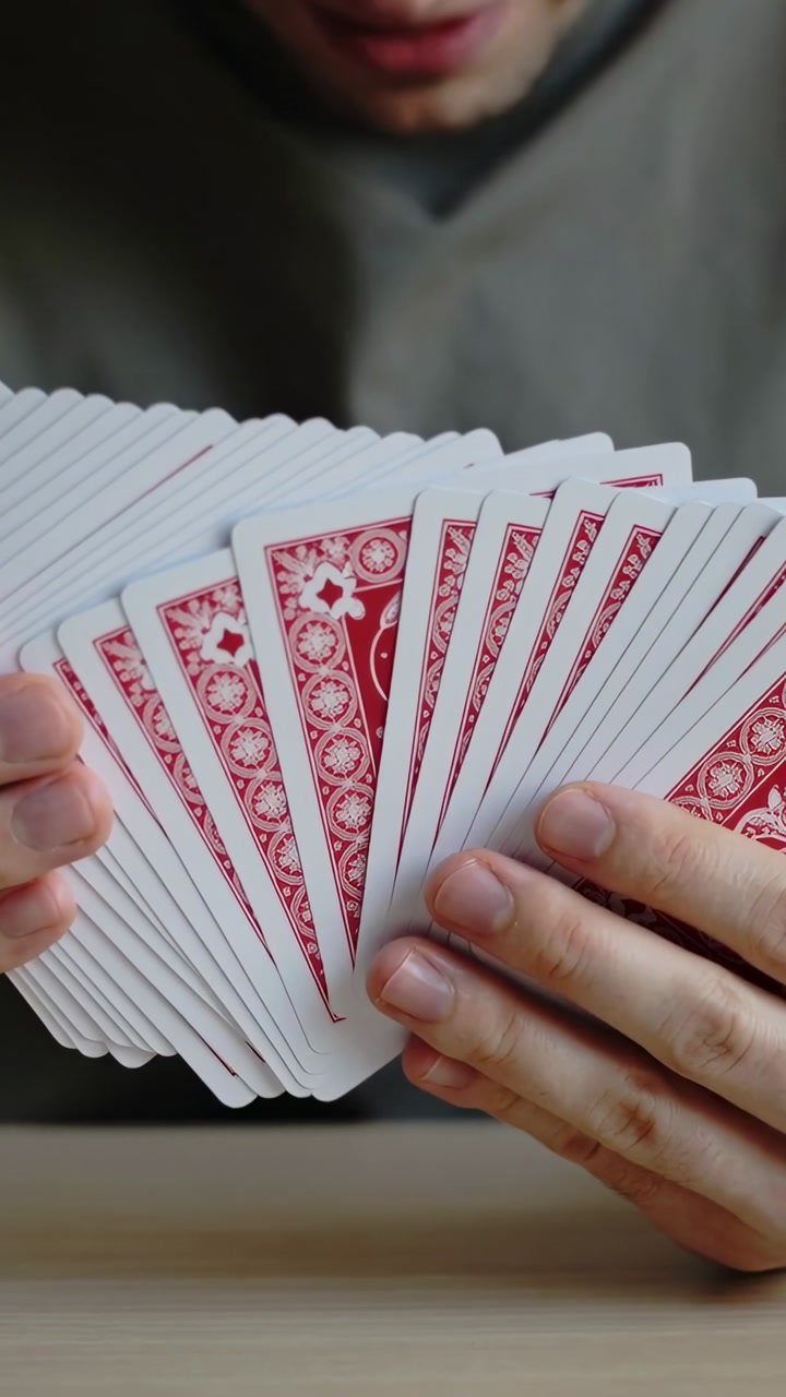 Vertical video: Holding deck, man fanning red-backed cards at wood table checking fan in grey shirt