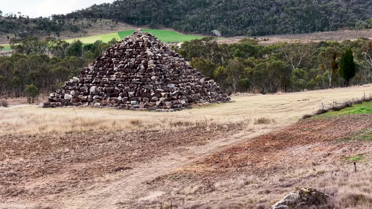 A dry stone pyramid stands in an open grassy field as the camera smoothly pans sideways, revealing the rural Australian landscape under soft daylight