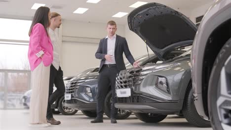 un joven vendedor de coches mostrando a una pareja de jóvenes un nuevo automóvil en el salón de concesionarios.