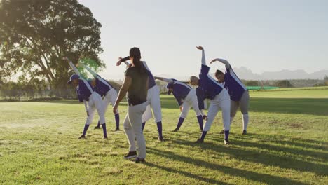 diverse group of female baseball players and coach on pitch, warming up, stretching from the waist