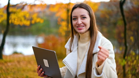 beautiful smiling lady holding a laptop speaking to the camera. dark-haired woman spending time in the nature in autumn.