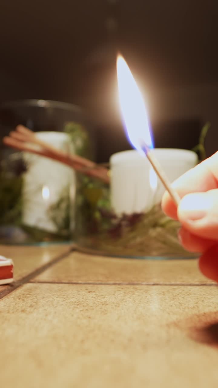 Tangan menyalakan korek api dan memegang api terbuka di dekat lilin di atas meja, menciptakan suasana rumah yang hangat dalam bidikan vertikal close-up