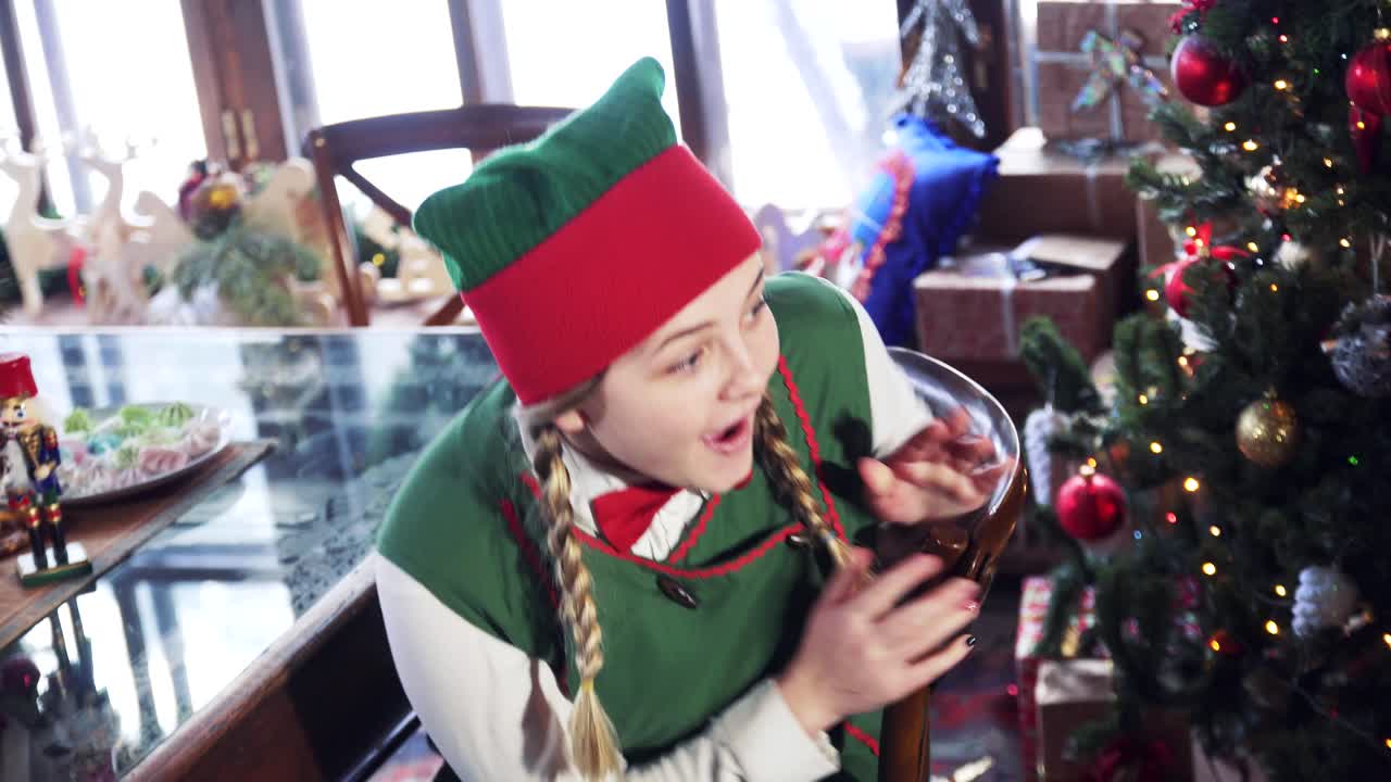 Excited elf sitting at the table. Joyful female in elf green costume is looking at something with surprise near the christmas tree