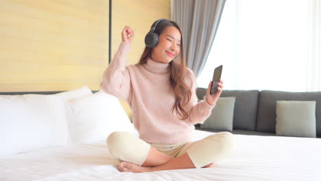 침대에 앉아 웃는 아시아 소녀는 헤드폰으로 음악을 듣습니다.