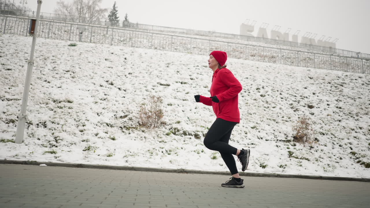 겨울 동안 눈 인 언덕 근처의 교차 도로에서 조하는 성인의 측면 뷰, 평온한 도시 환경, 벌거벗은 나무, 램프 기둥, 언덕 꼭대기에 보이는 디자인 글쓰기와 함께 빨간 후디를 입은 달리기 선수