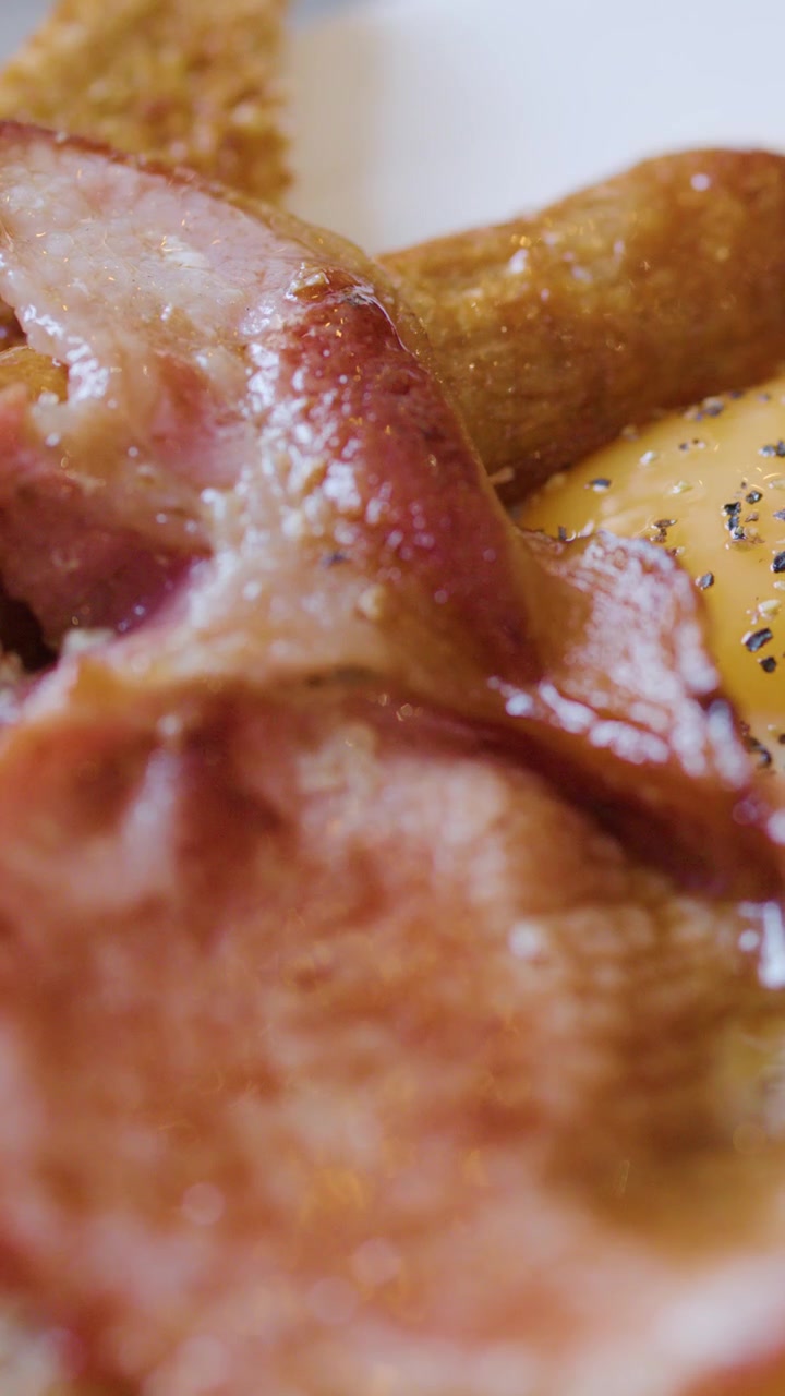 Una forchetta solleva salsiccia e pancetta da un piatto di colazione inglese con uova, toast, pepe, sotto luce naturale.