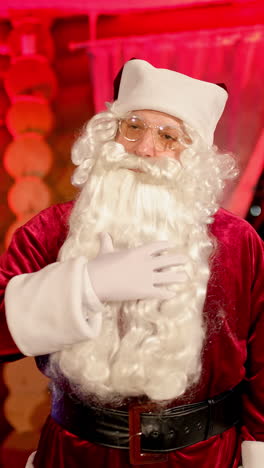 portrait of a senior man in santa claus costume indoors. serious santa with white beard standing in apartment on christmas tree background. christmas spirit. vertical video