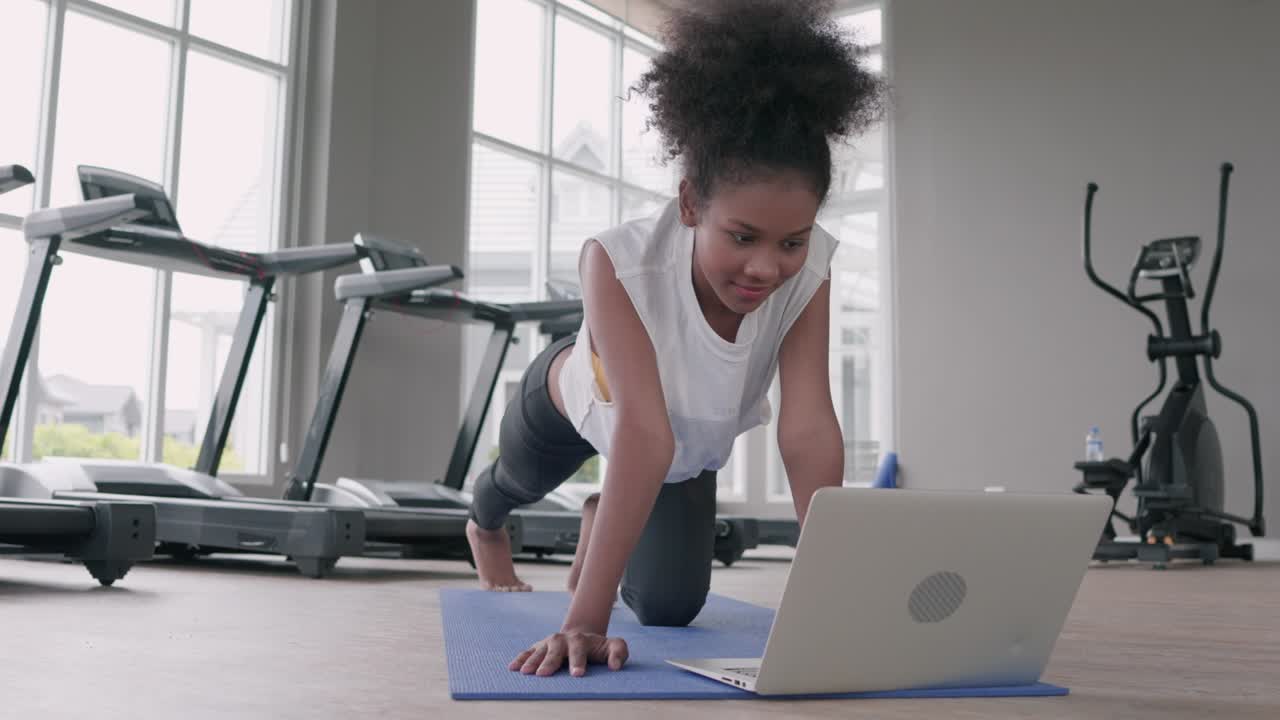 una mujer negra joven haciendo ejercicio de yoga en el gimnasio. una mujer afroamericana se ve en la lección de computadora portátil practicando la postura de tabla de yaga en la clase de internet en línea social.