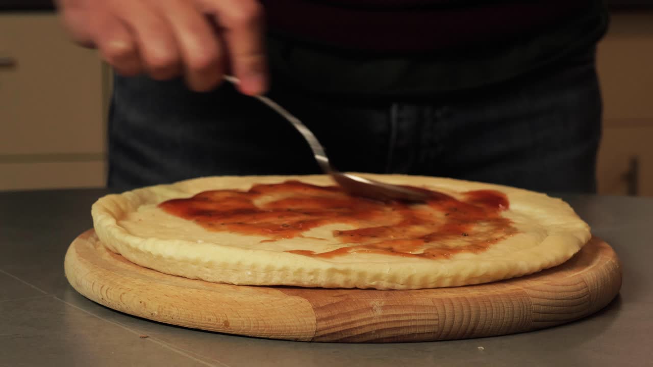 Un primo piano di una mano che spalma ketchup sull'impasto della pizza usando un cucchiaio, catturando le fasi iniziali della preparazione della pizza. Perfetto per illustrare la preparazione del cibo e i pasti fatti in casa