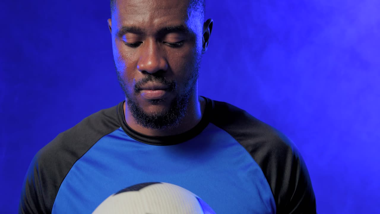 Uomo afroamericano che aggiusta la presa e abbassa un pallone da calcio, con una maglietta blu, in studio con inquadratura ravvicinata.