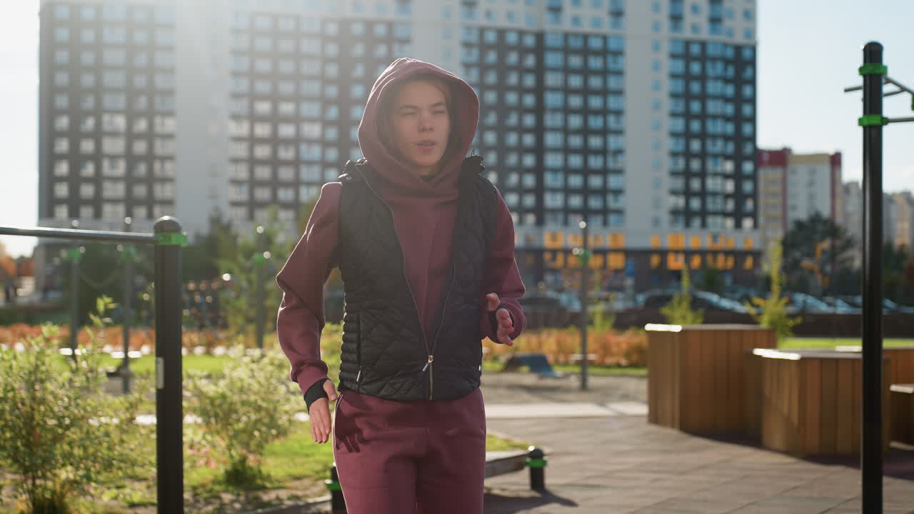 Strength trainer in hoodie and vest runs on spot raising arms and legs in urban street against apartments as pedestrians walk by under warm sunlight warming up for intense workout routine