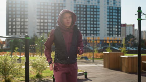 strength trainer in hoodie and vest runs on spot raising arms and legs in urban street against apartments as pedestrians walk by under warm sunlight warming up for intense workout routine